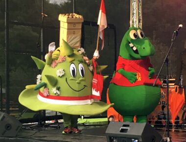 Das Kamenzer Stadtmaskottchen steht winkend mit Fähnchen auf einer Bühne neben dem Saurierpark-Maskottchen Bodo. Aufnahme vom Maskottchentreff beim Tag der Sachsen in Löbau als Beispiel für KAMI-Auftritte